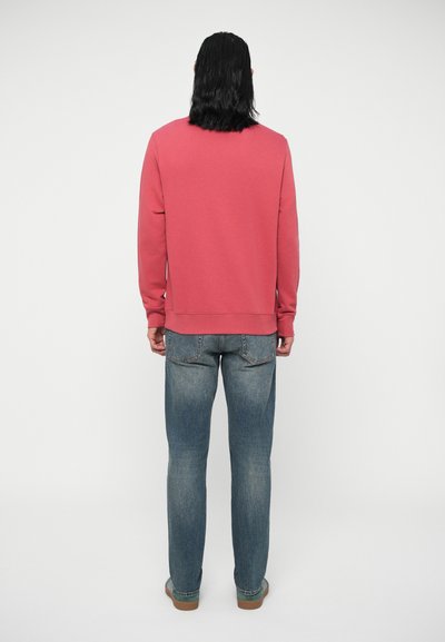 Asmuo su ilgais tamsiais plaukais stovi atsukęs nugarą, vilkintis rožinės spalvos džemperį, mėlynus džinsus ir laisvalaikio batus priešais šviesią, paprastą foną.