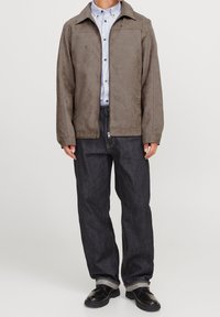 Brun ruskindsjakke med klassisk krave, frontlynlås og to sidelommer, parret med mørk denimjeans og sorte sko. Struktureret finish.