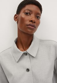 Manteau col classique gris clair en tissu texturé, comportant deux boutons foncés et une silhouette décontractée. Le fond uni met en valeur les détails.