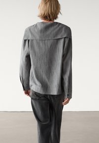 Persona con capelli corti biondi che indossa una maglietta a maniche lunghe grigia a righe verticali e jeans grigio scuri, girato di spalle su uno sfondo neutro.