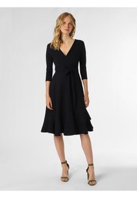 Schwarzes Wickelkleid mit tiefem V-Ausschnitt, dreiviertelärmeln und einem gestuften Rock. Merkmale sind eine gebundene Taille und es wird mit grünen Absatzsandalen kombiniert.