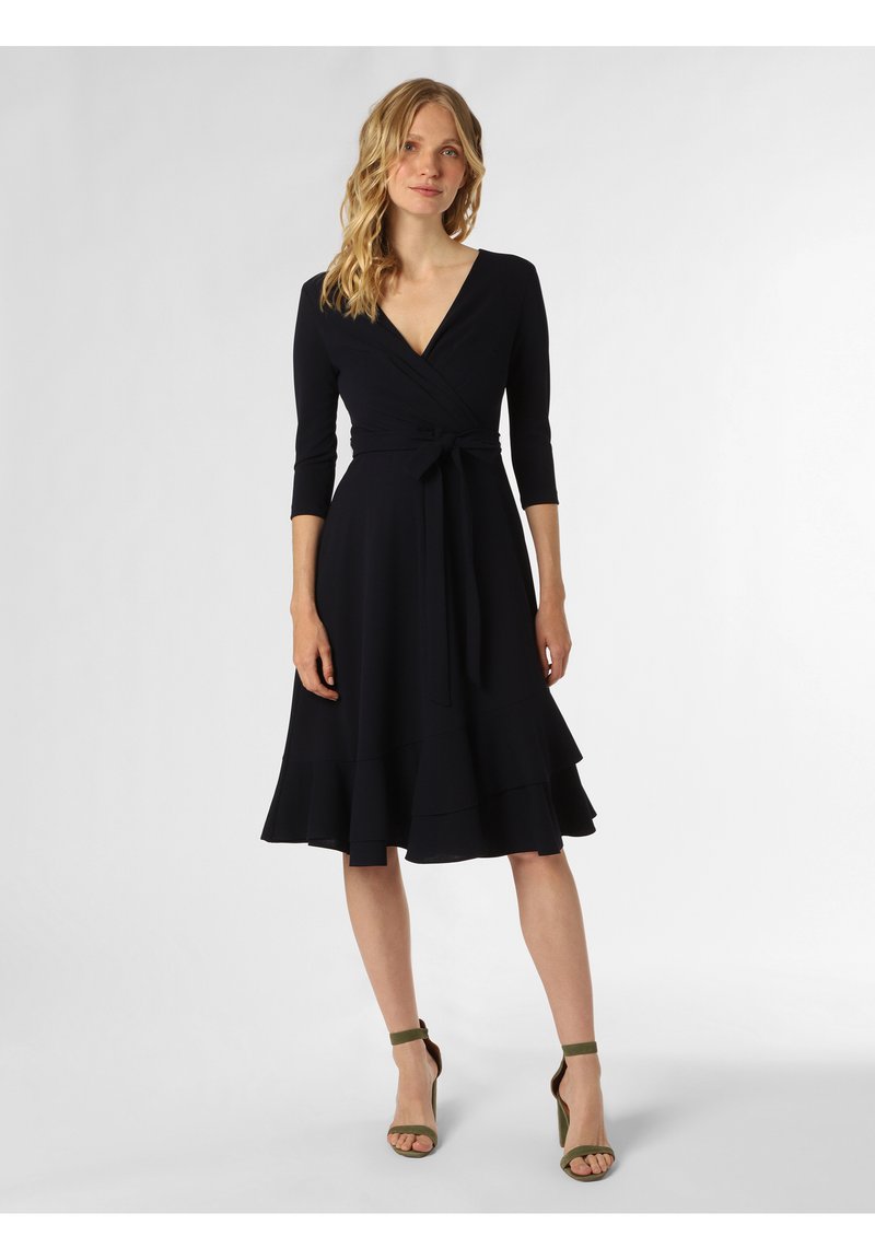 Schwarzes Wickelkleid mit tiefem V-Ausschnitt, dreiviertelärmeln und einem gestuften Rock. Merkmale sind eine gebundene Taille und es wird mit grünen Absatzsandalen kombiniert.