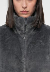 Femme portant une veste en fausse fourrure grise zippée avec un col haut et des cheveux bruns foncés lisses, vue de la mâchoire à la poitrine.