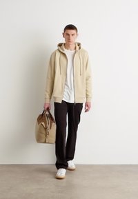 Felpa con zip beige sopra una t-shirt bianca, pantaloni neri e sneakers bianche. Porta una borsa beige con accenti marroni. Design minimalista.