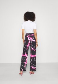Kvinna med lockigt hår, iklädd en vit t-shirt med korta ärmar, svarta byxor med ett klart rosa blixtmönster och rosa högklackade sandaler, stående.