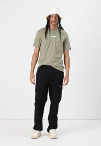 Khaki tričko s logem, spárované s černými cargo kalhotami s kapsami. Model nosí bílý klobouk typu bucket a světlé boty.