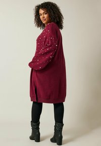 Cardigan oversize bordeaux avec de grandes accents en perles sur les manches, des poignets côtelés, des fentes sur les côtés et une coupe décontractée. Porté sur des leggings noirs et des bottes.