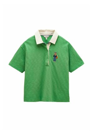 Zaļš īss piedurkņu polo krekls ar baltu apkakli un izšūtu lapsas tēlu, kas valkā zilus džinsus un tumšu jaku kreisajā krūšu pusē.