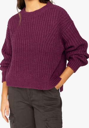 Kobieta w grubym fioletowym swetrze z dzianiny z prążkowanymi mankietami i dolnym brzegiem, w ciemnych spodniach typu cargo, z jedną ręką w kieszeni.