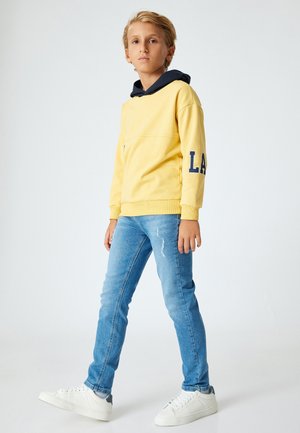 Felpa gialla con cappuccio navy e grafica "LA", abbinata a jeans chiari strappati e sneakers bianche. Look casual, moderno.