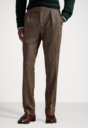 Homme portant un pantalon habillé à carreaux marron, des mocassins à franges marron foncé, un pull vert et une chemise bleue, les mains dans les poches, debout sur un sol uni.
