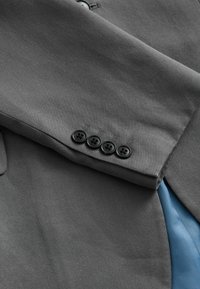 Graues Jackenmaterial mit glatter Textur, ausgestattet mit vier schwarzen Knöpfen am Ärmel und einer sichtbaren blauen Futter darunter.
