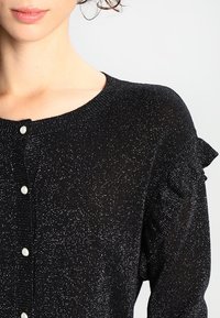 Svart stickad tröja med glittrande tråd, med rund halsringning, pärlknappar och volangdetaljer på axlarna.