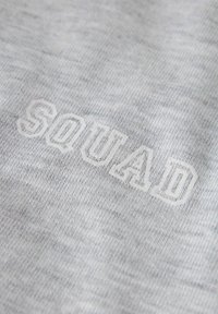 Close-up van lichtgrijze stof met wit omlijnde letters die "SQUAD" daarop geborduurd zijn.