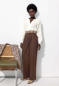 Chemisier blanc à boutons avec un tissu texturé, associé à un pantalon brun large taille haute et une ceinture beige clair. Boucles d'oreilles en or notables.