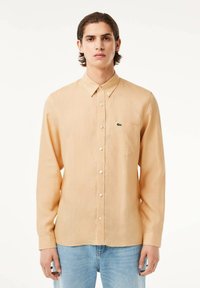 Camicia a maniche lunghe beige chiaro con colletto button-down, bottoni bianchi e un logo verde sulla tasca del petto. Tessuto morbido e vestibilità su misura.