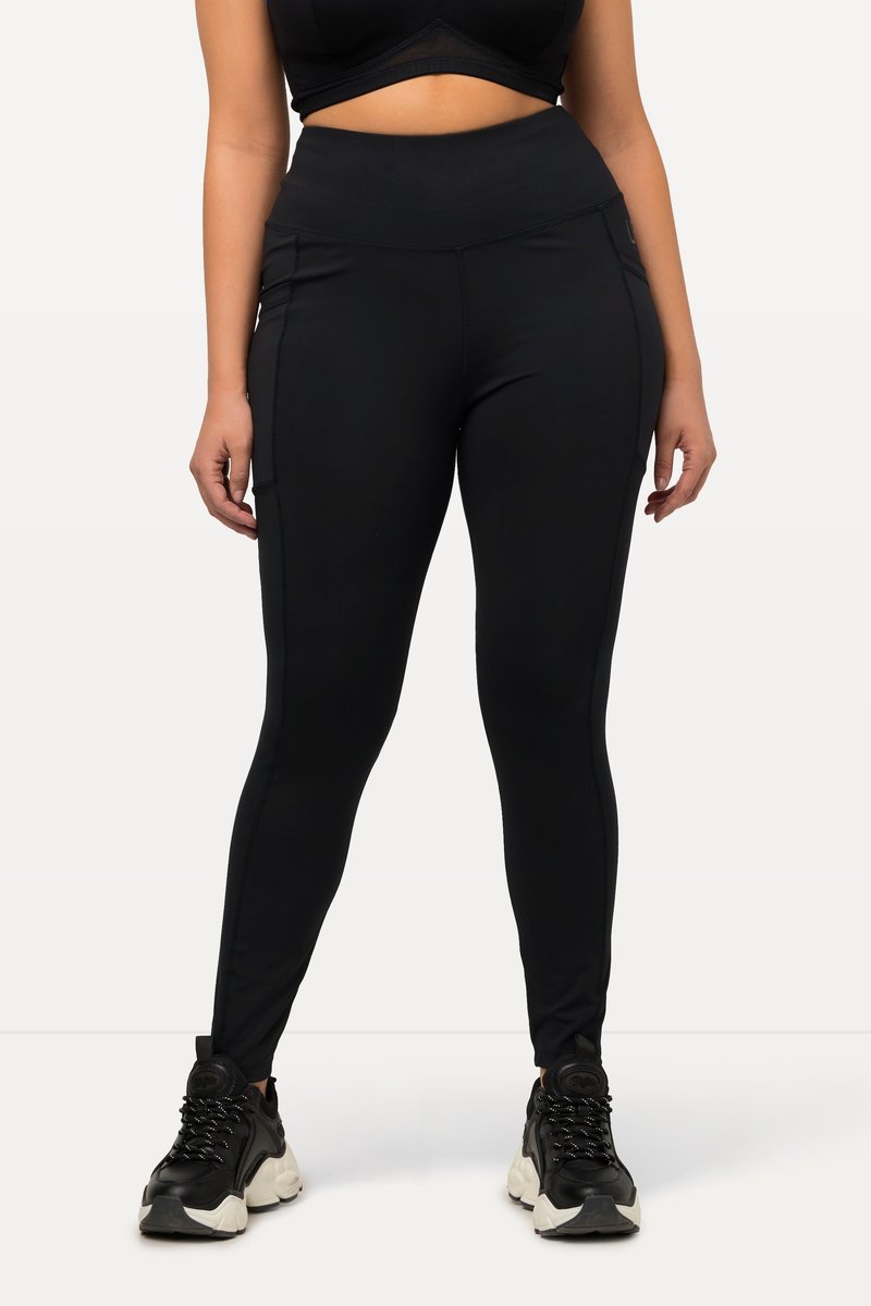 Frau trägt schwarze Leggings mit hohem Bund und seitlichen Taschen sowie schwarze klobige Sneakers, steht mit leicht gespreizten Beinen vor weißem Hintergrund.
