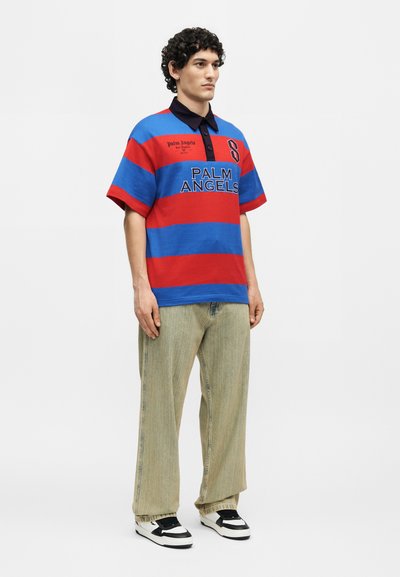 Νέος άνδρας φοράει μπλουζάκι πόλο Palm Angels με μπλε και κόκκινες ρίγες, χαλαρά μπεζ παντελόνια και ασπρόμαυρα αθλητικά παπούτσια, στέκεται μπροστά σε απλό φόντο.