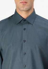 Chemise boutonnée en tissu teal foncé à motif à carreaux, avec un col structuré et cinq boutons noirs à l'avant.