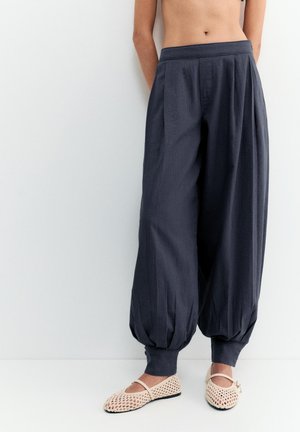 Osoba oblečená v voľných tmavomodrých nohaviciach s manžetami a béžových pletených balerínach, stojaca pred jednoduchým svetlým pozadím.