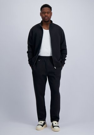 Mann steht mit den Händen in den Taschen, trägt eine schwarze Jacke mit Reißverschluss, ein weißes T-Shirt, schwarze Hose und schwarz-weiße Sneaker vor grauem Hintergrund.