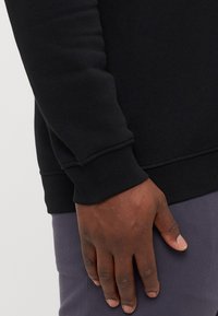 Black sweatshirt with ribbed cuffs and hem, featuring a soft texture. A hand rests on the hip, showing the garment's fit at the wrist.