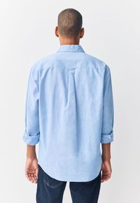 Lichtblauw overhemd met opgerolde mouwen, zachte textuur, afgeronde zoom en plooi detail aan de achterkant. Draagt bij donkere denim jeans.