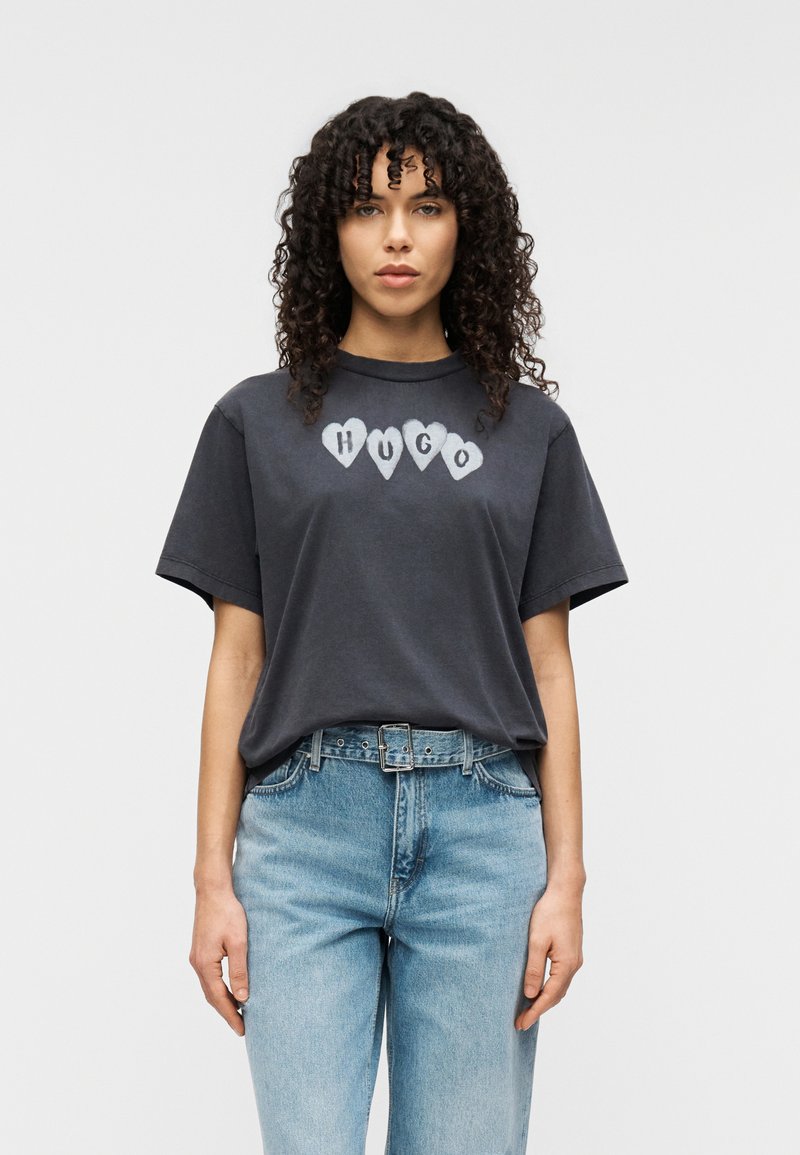 Σκούρο γκρι μπλουζάκι με στρογγυλή λαιμόκοψη και κοντά μανίκια, με λευκές καρδιές που έχουν το κείμενο "HUGO" στο κέντρο, συνδυασμένο με ανοιχτό μπλε τζιν.