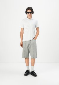 Polo de manga corta blanca con pequeños puntos negros, combinado con pantalones cortos de mezclilla gris claro. El modelo lleva gafas de sol negras, calcetines grises y zapatos negros.