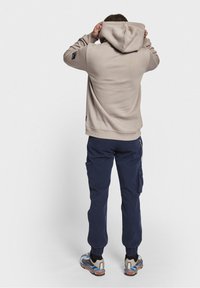 Beige hoodie with a drawstring hood and ribbed cuffs, paired with navy cargo pants. Notable detail includes a small logo patch on the sleeve.