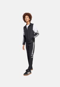 Zwart sportief trainingspak met witte strepen, bestaande uit een ritsjack en taps toelopende broek. Zwarte schoenen met bruine zolen maken de look compleet.