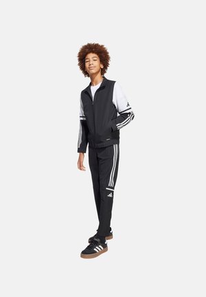 Completo sportivo nero con strisce bianche, composto da giacca con zip e pantaloni affusolati. Scarpe nere con suole marroni completano l'outfit.