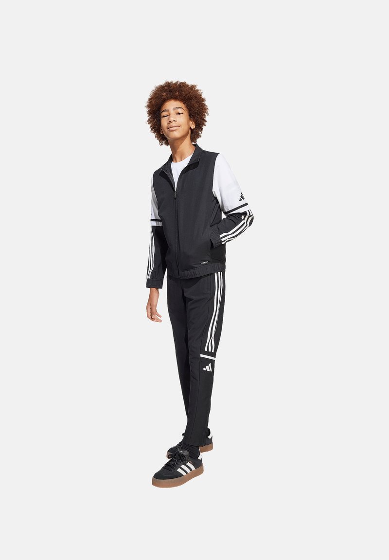Zwart sportief trainingspak met witte strepen, bestaande uit een ritsjack en taps toelopende broek. Zwarte schoenen met bruine zolen maken de look compleet.