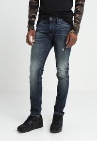 Jeans de denim azul de corte ajustado con un acabado desgastado y detalles rasgados. Combinados con zapatillas negras y una camisa de camuflaje de manga larga.