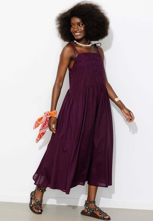 Femme avec coiffure afro portant une robe midi sans manches violet foncé, un collier coquillage, des sandales noires cloutées et une écharpe orange à motifs au poignet.