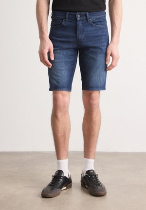 Donkerblauwe denim shorts met een nauwsluitend ontwerp, subtiele vervaging, vijf-pocketstijl en omgerolde zoomdetails. Gedragen met zwarte sneakers en witte sokken.