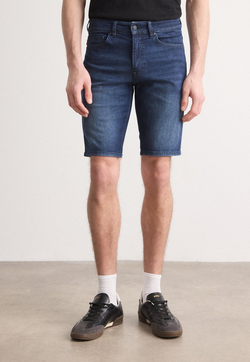 Shorts in denim blu scuro con un design aderente, lieve scoloritura, stile a cinque tasche e dettagli con orlo arrotolato. Indossati con sneakers nere e calze bianche.