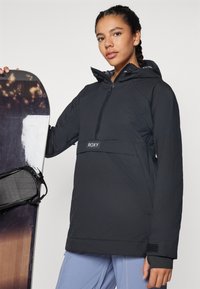 Black pullover jacket with a hood, featuring diamond quilted design, half zipper, and Roxy logo band; model holds a snowboard.