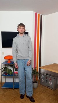 Νεαρός άνδρας με γκρι φούτερ και τζιν στέκεται σε έναν μοντέρνο χώρο με πολύχρωμες ρίγες στον τοίχο, πορτοκαλί φωτιστικό, φυτά και μια βαλίτσα με την επιγραφή "Tropicana".