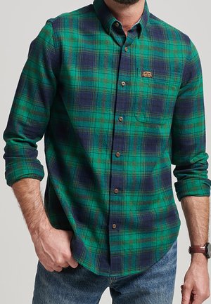 Mand iført en grøn og marineblå ternet skjorte med knapper, opkrogede ærmer, blå jeans og et brunt armbåndsur.