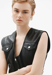 Chaleco de cuero negro con un escote en forma de V profundo, botones a presión y solapas en los hombros. Textura suave con costuras detalladas y sutil herrajes en plata.