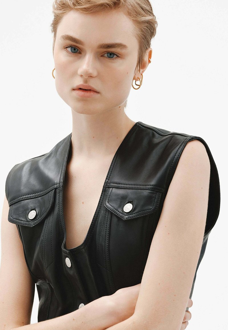 Chaleco de cuero negro con un escote en forma de V profundo, botones a presión y solapas en los hombros. Textura suave con costuras detalladas y sutil herrajes en plata.