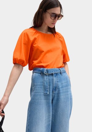 Kvinde iført solbriller med skildpaddemønster, lys orange puffærme-top, høj-taljede blå jeans med stofbælte, der holder en sort taske.
