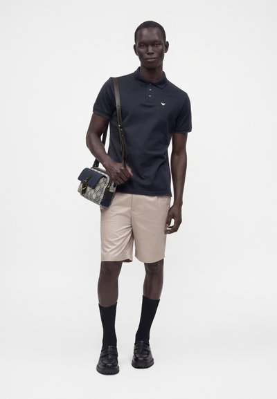 Homme portant un polo noir, un short beige, des chaussettes noires et des chaussures, portant un sac bandoulière à motif avec une sangle foncée sur l'épaule.