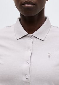 Gros plan d'une personne portant un polo gris clair avec un col, quatre boutons et un petit logo brodé sur la poitrine.