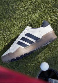 Λευκό και ναυτικό παπούτσι γκολφ που αιωρείται πάνω από τεχνητό γρασίδι κοντά σε μια μπάλα γκολφ που βρίσκεται στην άκρη μιας τρύπας.