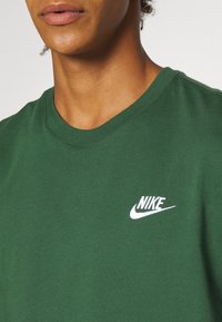 Detailní záběr na osobu, která má na sobě zelené tričko Nike s bílým logem Nike na levé hrudi.
