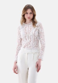 Top bianco in pizzo a maniche lunghe con scollatura e bordo scallop, decorato con modelli floreali, indossato sopra pantaloni color crema.