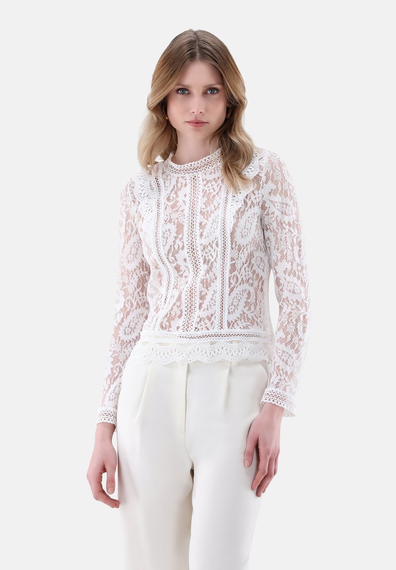 Top bianco in pizzo a maniche lunghe con scollatura e bordo scallop, decorato con modelli floreali, indossato sopra pantaloni color crema.