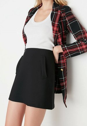 Kobieta w czerwono-czarnej kratowanej marynarce, białym topie na ramiączkach i czarnej, wysokiej spódnicy z przednimi klapkami kieszeni, stojąca na tle białej ściany.